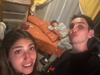 A selfie features three people indoors. A woman with long brown hair smiles at the camera. Beside her, a man with short dark hair poses with pursed lips. In the background, a person in a gray sweatshirt and pants rests on an orange mattress with patterned pillows. The setting appears casual, with draped fabric and shelves visible. Lighting suggests a cozy, informal environment.