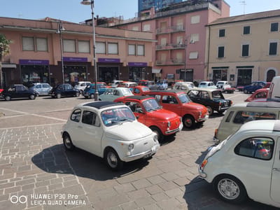 The image depicts a street scene in Follonica, Italy, featuring a gathering of classic Fiat 500 cars. The cars are parked on a cobblestone plaza in front of a row of buildings with shops. The buildings are in various shades of pink and tan, with awnings and store signs visible. In the foreground, a white Fiat 500 with a sunroof is prominently displayed, with the "HUAWEI P30 Pro LEICA QUAD CAMERA" watermark in the bottom left corner. Several other Fiat 500s, in colors such as red, black, and light blue, are parked behind it, creating a colorful display. Some of the cars have stickers and badges, including "Fiat 500 Club Italia." The sky is clear and blue, indicating a sunny day. The overall scene suggests a car enthusiast event or a local festival.