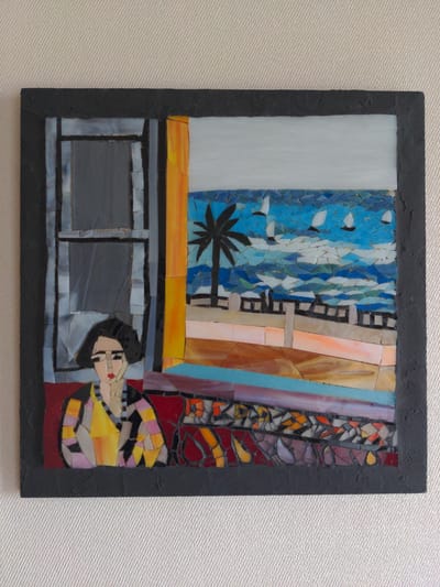 The image shows a framed mosaic artwork leaning against a textured wall. The artwork depicts a scene viewed through a window, with a woman on the left and a coastal landscape on the right. The woman, rendered in a stylized manner with dark hair and a yellow and black striped garment, is looking forward. Through the window, a palm tree stands against a blue sea with sailboats. A promenade or walkway is visible along the coast. The mosaic is made of small, irregularly shaped pieces of colored glass and other materials. The frame of the artwork is black and textured. The background wall is light-colored and has a woven texture. No specific location in Magherafelt, United Kingdom, is identifiable from the image.