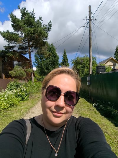 A selfie is taken by a woman with blonde hair, wearing large, dark sunglasses and a black shirt. She is outdoors in a rural setting with a dirt path in front of her. To her left is a wooden house and a brown fence, surrounded by lush green foliage and white wildflowers. To her right, a tall green fence runs along the path. In the background, a utility pole with numerous wires stands prominently against a partly cloudy blue sky. The overall scene suggests a village or countryside location, possibly in Новоникольское, Russia.