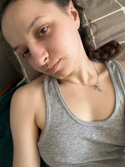 The image shows a close-up of a young woman lying down. She is wearing a gray tank top and a silver necklace with a cross pendant. Her hair is dark and curly, and it is pulled back to one side. She is lying on a bed with a patterned comforter. The lighting is natural and appears to be coming from a window. There is no visible indication of the city of Postoyata, Ukraine, in this image.