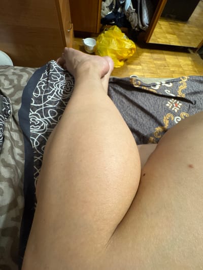 The image is an extreme close-up, low-angle shot of a person's lower legs and feet, lying on a bed. The person's left leg, particularly the calf, is prominently featured and appears muscular. The right leg is partially visible, showing the thigh and knee area. The bedding has a patterned design in dark blue, brown, and white. In the background, out of focus, is a wooden floor, a wooden dresser with drawers, and a yellow plastic bag. A white paper cup is also visible on the floor. There is no indication of the location being in the city of Лесные Поляны, Russia.