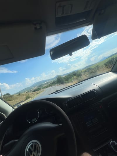 The image shows a view from inside a car, looking out through the windshield and toward the right side. The car's dashboard, steering wheel, and a portion of the center console with a radio are visible in the foreground. The steering wheel has a Volkswagen logo. Outside the car, a winding road leads through a hilly, rural landscape under a bright blue sky with scattered white clouds. The hills are covered with green vegetation and some patches of dry grass or crops. In the distance, there are more hills and mountains. The presence of a specific location in Svetlina, Bulgaria cannot be confirmed from the visual information alone. The scene appears to be a snapshot of a drive through the countryside on a sunny day.