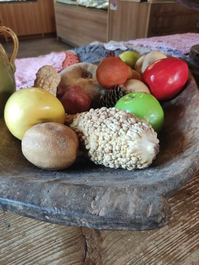 The scene, located in the city of Staraya Russa, Russia, presents a close-up, slightly elevated view of a weathered, dark wooden bowl filled with a still-life arrangement. The hand-carved bowl, exhibiting a rough texture and visible age, rests on a light-colored wooden table with distinct, parallel grain patterns.

Within the bowl, a collection of artificial fruits and natural decorative elements are arranged. Prominent items include a glossy yellow apple, a textured brown spherical object resembling a dried fruit or a small gourd, a distinctively bumpy, pale, elongated gourd with a visible stem, a shiny green apple, and a vibrant red apple. Additional items encompass an orange-colored spherical fruit, a reddish-brown irregular fruit, and a small, light green fruit. Several dark pinecones are interspersed among the fruits, alongside lighter-colored organic materials, possibly dried leaves or husks.

The background is significantly blurred, indicating an indoor environment. Visible details include indistinct wooden furniture or display cases and segments of various colored textiles, including pink, grey, and orange, suggesting decorative mats or coverings. No individuals or specific actions are depicted in the static arrangement.