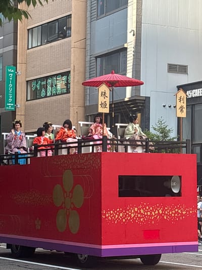 This detailed image captures a daytime scene on a city street in Kanazawa, Japan, likely during the Kanazawa Hyakumangoku Festival, given the traditional attire and names displayed. In the foreground, a large, rectangular parade float dominates the lower half of the frame. The float is primarily red with a purple base trim and is adorned with gold-colored decorative patterns, including prominent five-petal floral motifs and speckled designs. Two small wheels are visible beneath the float. On the right side of the float, a horizontal rectangular opening reveals a white megaphone speaker oriented outwards. Seated atop the float, behind a dark railing, are at least six individuals dressed in traditional Japanese garments (kimonos) and elaborate hairstyles. From left to right: a woman in a purple patterned kimono with an ornate traditional hairstyle is gesturing with her right hand; two young children in orange-red kimonos sit beside her; a woman in a lighter green kimono is positioned behind them; another person in an orange-red kimono is seated further right; a woman in a light green kimono with patterned lower garments (possibly hakama) holds an object; and a woman in a pink patterned kimono is partially visible on the far right. A large red traditional Japanese umbrella (wagasa) is mounted on the left side of the float, next to a wooden sign with the Japanese characters "珠姫" (Tamahime) written vertically in black. Another wooden sign, positioned further right on the float, displays "利常" (Toshitsune). The background features urban buildings. On the left, a multi-story building with light brown/beige horizontally ridged paneling is visible. On its second floor, people are visible through large windows, looking down at the street; one person wears a dark top. On the ground floor of this building, a large window displays green Japanese characters on a dark background, reading "ウィーククリニック" (Week Clinic), "心療内科" (Psychosomatic Internal Medicine), and "TEL". A vertical green sign attached to this building reads "Vie" and "ヴィーククリニック" (Vie Clinic) in white text. To the right, another building with lighter-colored (white/light grey) paneling features a small rectangular ventilation grate. At street level on the right, a black sign for "45 CAFE" is visible. A small green bush is situated between the float and the right-side building. The street surface is asphalt with a visible white line. The overall lighting is even, suggesting an overcast daytime. Faintly visible spectators in modern clothing can be seen on the far