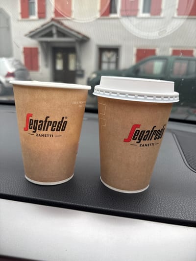 Two disposable coffee cups with the Segafredo Zanetti logo are placed on a textured black surface, likely the dashboard of a vehicle, with a silver-colored surface below. The cup on the left is open, while the cup on the right has a white plastic lid. In the background, a building with red shutters and a dark doorway is visible, along with the reflection of a person holding the camera and the front of a vehicle. The scene is situated outdoors, suggesting the vehicles are parked in a street or a parking area. The building in the background is consistent with architecture found in Untermünkheim, Germany.