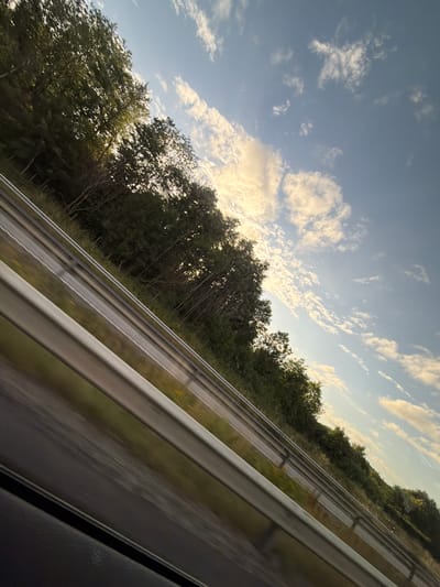 The image shows a view from a moving vehicle, likely on a highway, looking out towards a tree-lined embankment. The perspective is from a low angle, suggesting the camera is near ground level. The road has a metal guardrail in the foreground, which is blurred due to motion. Beyond the guardrail, the embankment is covered with dense green trees and vegetation. The sky above is blue with scattered white clouds, some of which are illuminated by the setting or rising sun, giving them a warm, golden hue. The image is tilted, adding a sense of movement. There are no people or specific landmarks visible that would definitively place it in Kungsbacka, Sweden. The scene is a generic highway landscape.