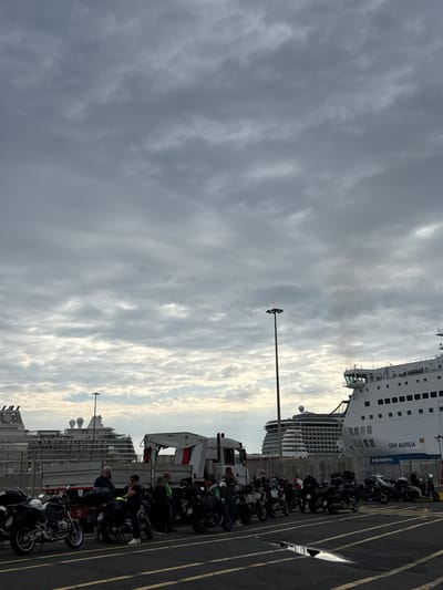 The image shows a parking area in Civitavecchia, Italy, adjacent to a large passenger ferry, the "GNV Auriga". A row of motorcycles are parked in marked spaces. Several people are standing near the motorcycles, appearing to be interacting or waiting. A white truck with a crane is positioned behind the motorcycles. The sky is overcast with clouds.
