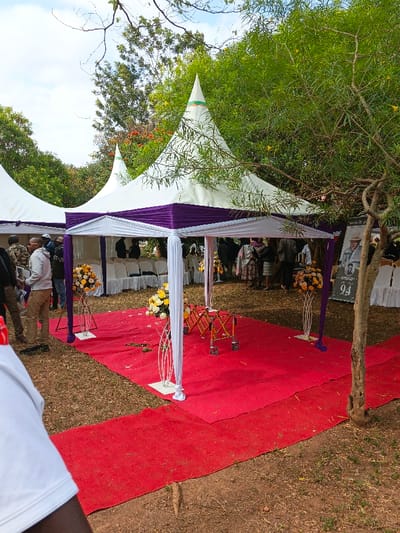 A red carpet leads to a canopy with white and purple trim. Inside, a collapsible stretcher rests on the carpet. Floral arrangements flank the canopy's entrance. White chairs are arranged within the canopy's covered area. A poster stands near a tree to the right, featuring a portrait and the number "94." People gather around the event. Additional white canopies are visible in the background, set amongst greenery.