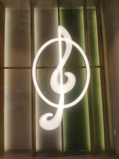 This media file shows a close-up, eye-level shot of a white, stylized musical note sculpture. The sculpture is illuminated and appears to be made of a smooth, possibly plastic or metal material. It is centered within a modern shelving unit. The shelving unit has multiple levels, with the sculpture positioned between two wooden shelves. The background behind the sculpture is a textured green surface, illuminated by hidden lights, which creates a gradient effect. Below the sculpture, the shelves are lit from above, casting a warm glow on the textured surfaces below. The overall impression is one of contemporary interior design, likely in a public space or a modern home. There is no visible text in the image, and no indication of the time of day or weather conditions. The scene is static and focused on the art piece and its display. The setting suggests an indoor environment, possibly a lounge area, a music room, or a decorative element in a restaurant or hotel.