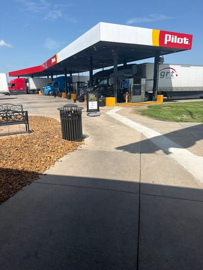 This is an eye-level, outdoor, medium shot of a Pilot gas station with multiple semi-trucks refueling under a covered canopy. The sky is clear and blue with a few wispy clouds. The Pilot logo, a red rectangle with a yellow arc and white text, is prominently displayed on the white canopy. Several fuel pumps are visible, with one black semi-truck hooked up and another blue semi-truck parked nearby. The ground is paved concrete with some areas of gravel and grass. A black metal trash can and a decorative metal bench are positioned on the gravel area in the foreground. The overall scene depicts a typical truck stop environment on a sunny day in Hayti, United States of America.