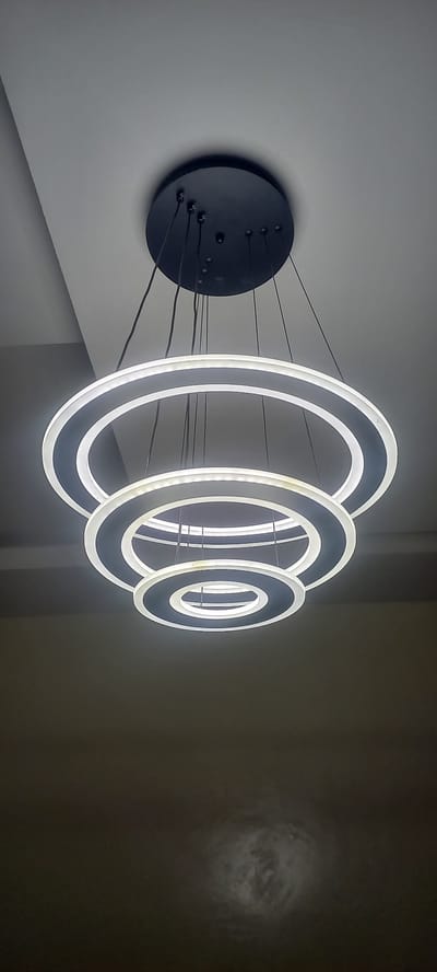 The image shows a modern, multi-tiered circular chandelier hanging from a ceiling. The chandelier consists of three concentric rings, each illuminated by LED lights around its inner circumference. The rings appear to be made of a dark material, possibly metal or acrylic, with a white inner edge where the lights are located. The chandelier is suspended by thin black wires from a circular black canopy mounted on a white ceiling. The lighting is dim, with the brightest illumination coming from the chandelier itself, casting soft light on the surrounding ceiling and a darker area below. The background is a plain, textured ceiling with a subtle gradient of light and shadow. There is no indication of any specific location in Laminga, Nigeria, or any people or actions.