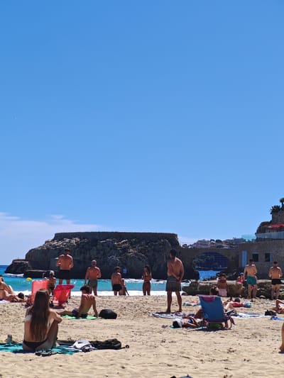 The photograph captures a beach scene on a sunny day. A group of people are enjoying various activities on the sandy shore, including sunbathing, standing, and interacting near the water's edge. In the background, there is a rocky headland with a built structure on top, and a bridge connecting it to another structure. These structures are likely part of the cityscape of Sant Agustí, Spain. The sky is clear blue, and the sea is visible between the people and the headland. There are various beach chairs and towels scattered on the sand.