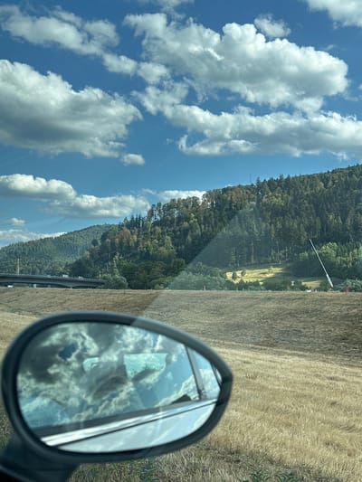 An outdoor, eye-level, vertical, medium shot captures a landscape scene from within a vehicle, as indicated by the reflection in the side-view mirror. The mirror itself, in the foreground and on the left, reflects a blue sky filled with fluffy white clouds.

Beyond the mirror, the scene unfolds to reveal a vast expanse of dry, golden grass covering rolling hills. In the mid-ground, a highway overpass, carrying vehicles, stretches across the landscape. Further back, a densely forested hill rises, with trees of varying shades of green, some showing hints of autumnal color. A tall, white, crane-like structure is visible on the right side of the forested hill. The sky above is a vibrant blue, dotted with scattered white clouds.

Based on the available information, this scene is located in the city of Haslach im Kinzigtal, Germany.