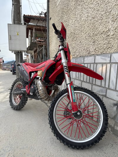 A red GasGas enduro motorcycle is prominently parked on a paved surface, likely a sidewalk or street, in Breznitsa, Bulgaria. The motorcycle is positioned facing forward and slightly to the right, leaning on a kickstand which is not visible but implied by its upright posture. It features red plastic bodywork, including the front fender, tank shrouds, and seat. The front wheel has a silver rim with distinct red spoke covers, and is fitted with a knobby tire. The front suspension forks are silver, displaying "WP" and "MOTOREX" branding. A protective metal cage surrounds the exhaust pipe. The motorcycle's engine area is dark-colored.
To the right, a textured, light-colored building wall, possibly stucco or rough plaster, forms the immediate background, with a lower section of horizontal light grey blocks. To the left and rear of the motorcycle, a grey utility pole with a white electrical box attached is visible. Beyond the pole, an older residential building with exposed wooden elements, a balcony, and bare vine growth is present. Further down the sloping street, a dark-colored SUV or truck is parked. In the distant background, rolling hills or mountains are visible under an overcast sky. No individuals are present in the scene, and no specific actions or interactions are depicted beyond the motorcycle being stationary.