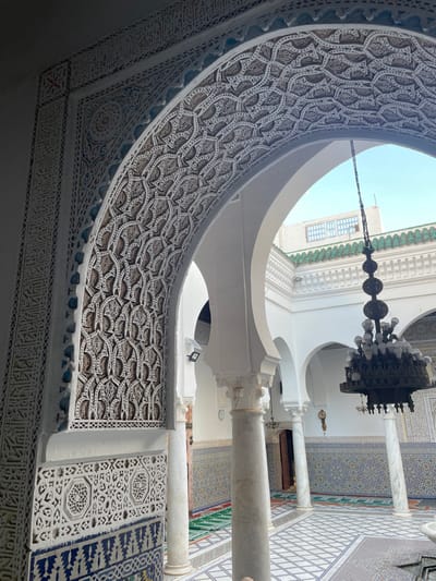 The image depicts an arcaded courtyard or hall within a religious or historical complex, identified as the Mausoleum of Moulay Idriss I in Moulay Idriss Zerhoun, Morocco. The foreground prominently features an elaborate horseshoe arch adorned with intricate white stucco (gesp) carvings featuring geometric patterns, floral motifs, and calligraphic script, with dark brown accents. The lower section of the wall beneath this arch is decorated with blue and white geometric mosaic tiles (zellij).
Beyond this arch, an open courtyard is visible, surrounded by white plastered walls and multiple supporting white horseshoe arches, resting on white marble columns with fluted shafts and decorative capitals. The courtyard walls are extensively covered with colorful geometric mosaic tilework, predominantly in blue, white, and yellow, extending to a mid-wall height. The courtyard floor is paved with patterned square tiles.
A large, multi-tiered, dark metal chandelier with numerous white light bulbs hangs centrally within the visible courtyard space. A section of a white marble fountain is discernible in the lower right foreground of the courtyard. On the distant walls, a pendulum wall clock is visible. Portions of a green-tiled roofline, characteristic of Moroccan architecture, are seen in the upper background, against a clear blue sky, indicating an open-air environment and daylight illumination. Several green prayer mats are laid out on the tiled courtyard floor. No individuals are present within the scene.