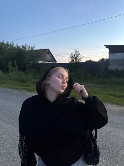 A young woman with blonde hair, wearing a black hooded sweatshirt, stands outdoors on a paved surface, possibly a road, in the village of Ryabinino, Russia. She is leaning her head back and puckering her lips as if for a kiss, while her right hand adjusts the hood of her sweatshirt. A black handbag hangs on her right side. The background features a wooden fence, vegetation, and a rustic building with a peaked roof, indicative of a rural or semi-rural setting. The sky above is a clear blue with some wispy clouds, suggesting it is either early morning or late evening.