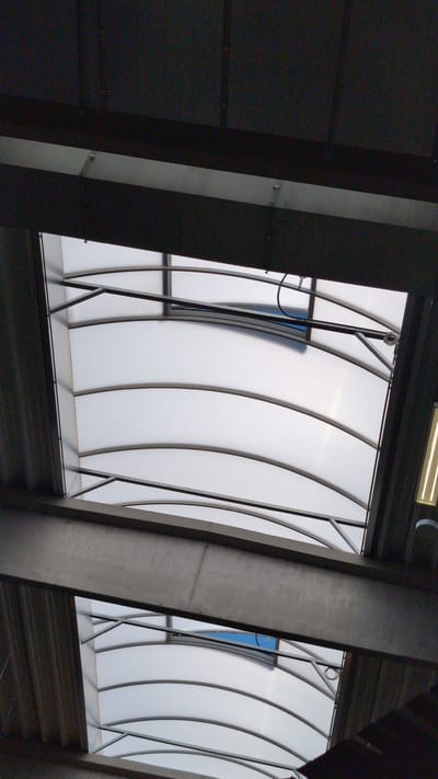 This media file captures an interior view of a modern architectural space, likely a building or a transit hub, in Vysoké Mýto, Czechia. The dominant subjects are structural elements of the building. Large, dark grey concrete columns flank the scene, creating a sense of enclosed space. Between these columns, a translucent, curved ceiling or wall is visible, composed of white, opaque panels framed by dark, arched ribs. This unique design gives the ceiling a flowing, organic appearance, reminiscent of waves or ribs.
Through gaps in the translucent panels, glimpses of a bright blue sky are visible, suggesting that this is an outdoor-facing element, possibly a roof or a large window structure. The lighting suggests it is daytime, with a long, fluorescent light fixture visible at the top of the frame, indicating artificial illumination within the structure.
The overall impression is one of modern, functional architecture, with a focus on light and form. There are no people or specific activities depicted. The scene is static, focusing on the architectural details and the interplay of light and shadow. The concrete textures of the columns and the smooth, curved surfaces of the ceiling create a contrast in materials. The metal framing and supports for the curved panels are visible, showcasing the construction. No visible text is present in the image.