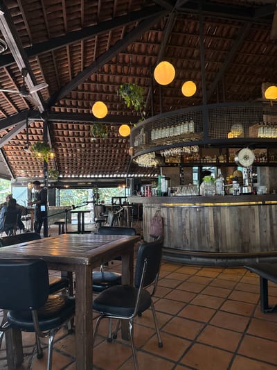 The image shows the interior of a restaurant, featuring a bar area and dining tables. The roof has a distinctive design with terracotta tiles and exposed black metal support beams. Globe-shaped light fixtures hang from the ceiling. A circular bar with a wooden facade is visible, along with shelves displaying glassware and bottles. People are seated at tables in the distance. The floor is tiled with terracotta-colored squares.