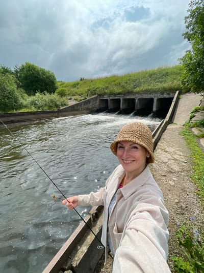 A woman wearing a straw hat and a light-colored jacket smiles at the camera while fishing on a concrete embankment next to a body of water. The water appears to be flowing from a series of concrete culverts in the background, suggesting a dam or spillway. The surrounding environment is overgrown with green grass and trees. The sky is cloudy. The location is in Bereznik, Russia.