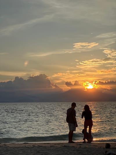 A group of people are silhouetted on a sandy beach facing a body of water during sunset. The sky is filled with colorful clouds, with the sun partially obscured behind them, casting a warm glow. The water is calm with gentle waves lapping at the shore. The people appear to be enjoying the view, with two standing and one crouching on the sand. The scene captures a peaceful moment at the beach, likely in Saavedra, Philippines, given the tropical setting and common tourist activities.