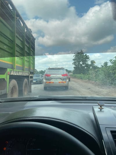 The image shows a view from inside a vehicle, likely a car, looking forward through the windshield. The scene appears to be a traffic jam on a dusty, unpaved road in a rural or semi-rural area. In the foreground, the side of a large green truck with yellow accents is visible. The truck has large tires and the text "ALLAH PENUH" is visible on its side in white lettering. Further ahead on the road, a light-colored SUV with tinted windows is stopped. The rear of the SUV shows taillights and some writing on the rear window, but it is largely unreadable. Behind the SUV, more vehicles are visible, indicating a queue. The road is flanked by lush green vegetation and trees. The sky is overcast with dramatic, billowy clouds, suggesting a humid or possibly stormy day. The overall lighting indicates it is daytime. A small circular magnetic phone mount is attached to the dashboard of the car from which the photo is taken. The dashboard itself is dark gray plastic. On the right side of the image, part of the car's instrument panel is visible, with a speedometer showing readings around 40, 60, and 80, and some indicator lights illuminated. The scene implies a pause or slow movement in traffic on a road in Aciol, Nigeria.