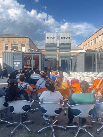 The image depicts an outdoor gathering of people seated on office chairs, facing a row of shipping containers repurposed into a venue. The scene is set in Gladzor, Armenia. The weather appears to be partly cloudy with a bright blue sky, suggesting a daytime event, likely in the late morning or afternoon.

The subjects are a diverse group of individuals, with a focus on their backs as they are seated and facing away from the camera. They are of various ages and genders, dressed in casual attire. The chairs are an eclectic mix of white, orange, and red, with chrome bases, contributing to a somewhat informal and perhaps temporary setup.

The shipping containers form a backdrop, with some open to reveal interior spaces, possibly used for exhibitions or presentations. Two large, white signs with dark lettering are visible on the containers. One sign contains the word "rankun" in Armenian, and the other has an indistinguishable logo. The overall atmosphere seems relaxed and communal, possibly an outdoor workshop, a lecture, or a social gathering. The ground is paved with asphalt or gravel, and there is some greenery visible on the right side of the frame. The architecture of a building with windows is visible in the background, hinting at an urban or semi-urban environment. The image is oriented vertically, making the scene appear foreshortened.