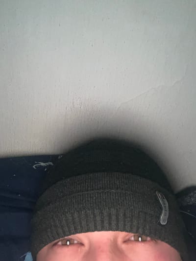 A close-up image shows a person wearing a dark green knitted beanie. The beanie has a small, metallic logo. The person's eyes are visible and appear to be brown. Part of their nose and mouth can be seen. The background is a plain white wall. The lower part of the image includes a blue shirt and a few white markings. The image appears to be rotated.