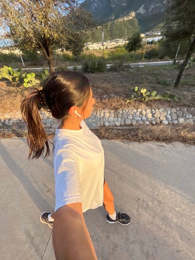 A woman in a white t-shirt and black shorts walks along a concrete path. She wears black sneakers and white socks. Her dark hair is pulled back in a ponytail. She wears white wireless earbuds. The path is bordered by a stone wall and greenery. Buildings and a mountain are visible in the background. The sunlight casts long shadows.