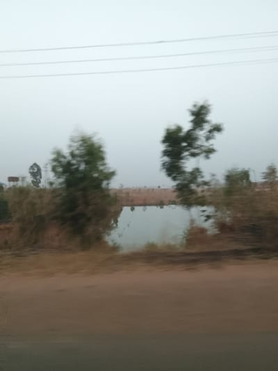 This is a blurry, overcast, outdoor image taken in Rahwol Chom, Nigeria, likely from a moving vehicle. The foreground features a wide, reddish-brown ground surface, possibly an unpaved road or dirt shoulder, extending across the lower portion of the frame. In the midground, a calm body of water, appearing as a pond or lake, reflects the light grey sky. The water's edges are obscured by blurred green and brown vegetation, including several distinct trees. The background reveals a reddish-brown elevated terrain or embankment beyond the water, with several indistinct structures on the distant horizon. One prominent background object appears to be a tall, metallic tower, possibly a water tower or cell tower, to the left. Multiple dark, horizontal power lines traverse the upper sky against the hazy, uniformly light grey backdrop. No individuals or specific actions are discernible.