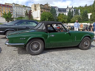 The image depicts an outdoor scene in Mariánské Lázně, Czechia, with a focus on a vintage green Triumph Spitfire IV convertible parked on a textured grey surface, possibly a street or parking lot. The car's black soft top is partially deployed. Behind the car, a dark-colored coupe is visible.

In the background, several buildings with varying architectural styles and colors line a street. Some buildings appear to be older, with ornate facades and balconies, while others have more modern or weathered appearances. The buildings are set against a backdrop of lush green trees and a cloudy sky.

Several people are scattered throughout the scene. In the mid-ground, a man in a light blue shirt and white pants stands near a group of people. Other individuals are seen walking or standing near parked vehicles, suggesting a gathering or event, possibly a car show. The overall impression is a pleasant day with classic cars displayed in a picturesque European town.
