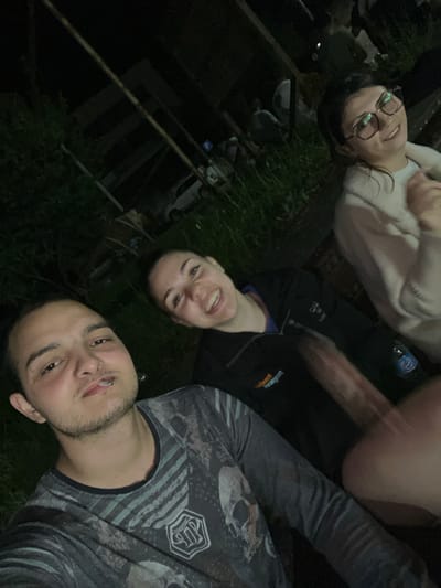 In an outdoor, nighttime setting, three individuals are captured in a selfie. In the foreground, a man with short dark hair and a faint beard smiles towards the camera, holding a cigarette between his lips. He is wearing a dark grey t-shirt with a skull graphic and horizontal stripes. To his right, slightly behind him, a woman with dark hair and a wide smile looks towards the camera. She is wearing a black jacket over a blue undershirt. Further in the background, a woman with dark hair and glasses smiles and looks slightly away from the camera. She is wearing a light-colored sweater. The environment appears to be a grassy area with some vegetation and what looks like wooden structures or fences in the background, hinting at an outdoor gathering or event. The overall lighting suggests it is dark, with artificial lights illuminating the subjects. The scene is likely in Dragoevo, Bulgaria, given the context provided.