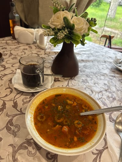 A table set with a patterned tablecloth, a bowl of soup, a glass of dark liquid, a vase of artificial flowers, and a bottle of amber liquid. The soup is a rich red color with visible pieces of ingredients and green herbs. The glass contains a dark beverage, likely coffee or tea, and rests on a saucer with a folded napkin and a spoon. A brown vase with white artificial roses and green leaves is positioned behind the glass. A tall glass bottle filled with amber liquid, possibly oil or vinegar, is partially visible on the left side of the frame. The background suggests an indoor setting with a window showing greenery and a blurred outdoor scene. The overall impression is a meal at a table, possibly in a restaurant or home. The image does not provide any specific details about the city of Bolotnitsa, Russia.