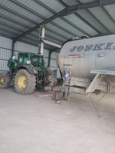 The image shows a green John Deere tractor coupled to a silver Joskin agricultural tanker inside a building with a metal frame and corrugated metal siding. The tractor has the license plate "1.XEX 1741," suggesting it's registered in Bulgaria. The scene is likely in a rural or agricultural setting. The location could be Ovcharovo, Bulgaria, as requested, but there are no specific location markers within the image to confirm.