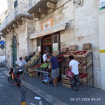 The image shows a fruit stand on a city street in Ruvo di Puglia, Italy. The fruit stand is set up in front of a building with stone facade and a balcony. There are several people present. An older man wearing a white striped shirt, light-colored shorts, and a white cap stands near a scooter. Another man, wearing a blue t-shirt and gray shorts, is examining produce at the stand. A person, whose face is not visible, wearing a white t-shirt and floral patterned pants, is shopping at the stand and holding a bag. The fruit stand displays a variety of fruits and vegetables, including melons, tomatoes, peaches, and what appear to be gourds or large green pears. Wooden crates are used to hold the produce, and price signs are visible. An orange scooter is parked on the sidewalk. The environment appears to be a sunny day. The street is paved, and there is a curb and sidewalk. A blue pedestrian crossing sign is visible on a pole. The building above the fruit stand has an awning and a sign that reads "La Frutteria da Angelo Ficco".