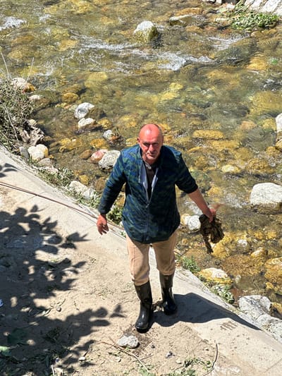 A man with a bald head stands on a concrete embankment next to a shallow, rocky stream. He is wearing a blue and green patterned jacket, tan pants, and black rubber boots. In his right hand, he holds a dark, dirty piece of fabric. The stream bed is filled with yellow and brown rocks, and the water is clear and flowing. Green vegetation grows along the bank of the stream and on the embankment. Shadows of leaves are cast on the concrete. The location is the city of Dulovine, Montenegro.