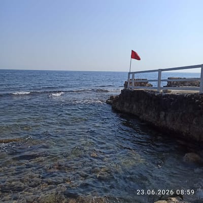 The image captures a serene coastal scene with the clear blue sea meeting a rocky shoreline. A white railing runs along the top of the rocky edge, suggesting a walkway or viewing platform. A bright red flag is prominently displayed, attached to a pole extending from the railing, likely indicating a warning or advisory for swimmers. The water is calm, with gentle waves lapping against the shore. Sunlight reflects off the water's surface, hinting at a clear day. The timestamp in the upper right corner indicates the image was taken on June 23, 2026, at 8:59 AM, suggesting it's early morning. The presence of the flag and the calm sea points towards a potentially safe but possibly regulated area for water activities. The rocky terrain and the railing suggest a developed coastal area, possibly a public beach or a scenic overlook.