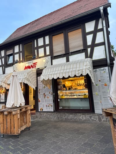 The image captures the exterior of "EISMANUFAKTUR MONTI," an ice cream shop located in Bruchköbel, Germany, on an overcast day. The building showcases traditional half-timbered architecture with dark brown beams contrasting with white and green-bordered infill panels. A reddish-brown tiled roof and a metal rain gutter are visible at the top.
The shopfront features a large display window and a glass door, revealing illuminated display cases inside, likely containing ice cream or pastries. Two white and beige striped awnings are extended above the window, providing shade, while another is folded. Large menu boards are prominently displayed next to the entrance, listing a variety of offerings in German.
Key menu categories include "Caffè & Drinks" (coffee and drinks), "TEE" (tea), "KAKAO" (cocoa), "ICED CAFE," "EISTEE" (iced tea), "MATCH," "WASSER" (water), "LIMO" (lemonade), "APERITIVO" (alcoholic and non-alcoholic aperitifs), "SMOOTHIE," and "Protein & Greens." Further boards detail "KLASSIKER" (classics), "FRUCHT EISBECHER" (fruit sundaes), "SCHOKOLADIG" (chocolate sundaes), "MIT ALKOHOL" (with alcohol), "UNSERE FAVORITEN" (our favorites), "BRIOCHE," "EISKAFFEE 6,50" (iced coffee for €6.50), "SOSSE 1,00" (sauce for €1.00), "SPAGHETTI LIEBE" (spaghetti ice cream), "KINDER HIGHLIGHTS" (children's highlights), and "EISGETRÄNKE" (ice cream drinks). Prices, denominated in Euros, are visible for many items, such as "Aperol Spritz 6,50 €" and "Erdbeer Spaghetti 8,50 €." A sign below the window reads "FOLLOW US ON Instagram @eiscafe_monti_bruchkoebel."
In front of the shop, the ground is paved with dark grey cobblestones, with a section of natural stone cladding along the building's base. Several wooden pallets, some with "EPAL" branding and equipped with wheels, are arranged near folded white outdoor umbrellas, suggesting outdoor seating or display. No people are visible, but the open shop and outdoor setup indicate business is active.