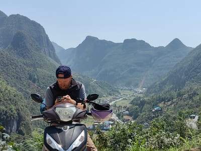A man is sitting on a parked moped in front of a scenic mountain landscape in the Phố Bảng Commune, Vietnam. The man is fair-skinned, has a beard, and is wearing a blue baseball cap, gray shirt, and black vest. He is looking down at a phone in his hands. The moped is black and gray with a purple helmet hanging on the handlebar. In the background, rolling green mountains stretch into the distance, with a small town visible in the valley below. The sky is clear and blue. Vegetation is visible along the roadside.