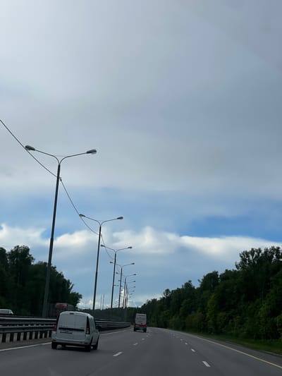 The image depicts a highway scene in or near Бавыкино, Russia, on a cloudy day. The view is oriented along the road, showing a line of lampposts alongside the highway. A white van is driving on the right lane, closer to the camera, with another vehicle visible further ahead. Trees border the highway on the right side. The sky is predominantly covered with clouds, showing some patches of blue.