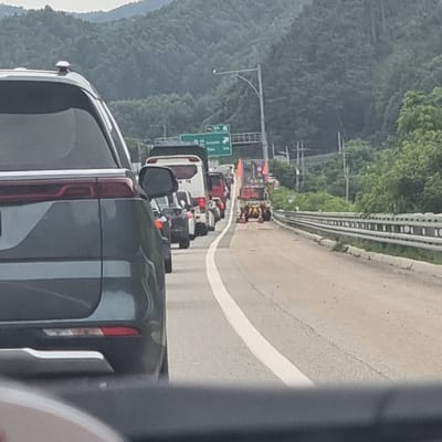 The image depicts a traffic jam on a highway in Jinbu-myeon, South Korea. In the foreground, a dark-colored SUV is stationary, its rear visible. Ahead, a line of vehicles, including a white bus, a red van, and several other cars and trucks, are also stopped. Further down the road, construction equipment, including a yellow and orange vehicle with cones, is present, suggesting potential road work or an accident. The highway is bordered by a guardrail and dense, tree-covered hills in the background, indicative of a rural or mountainous setting. The sky is overcast, and the overall atmosphere suggests a humid, summer day. There are no people clearly visible in the scene.