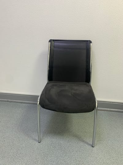 An empty, black mesh-backed chair with a black upholstered seat and chrome legs sits in front of a textured white wall with a grey baseboard. The floor is a speckled grey linoleum. The scene is indoors, possibly an office or waiting room. No people are present. The image is taken from a frontal perspective, slightly angled down.