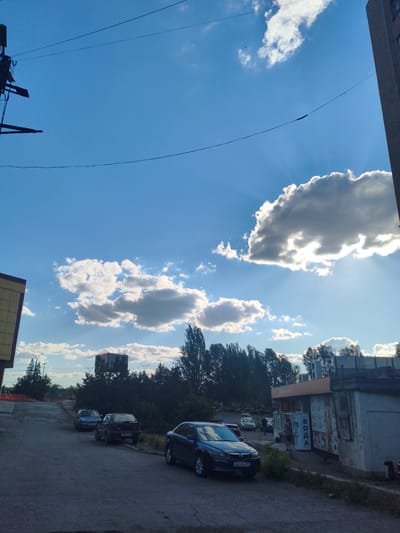 This image depicts a street scene in Alchevsk, Ukraine, under a bright, partly cloudy sky, suggesting it is daytime, likely late afternoon given the angle of the sun. A large, dark cloud is prominently featured, with sunlight breaking through its edges, creating visible sunbeams. In the foreground, a dark blue car is parked on the right side of the asphalt road, with its license plate visible. To the left, another dark-colored car is parked, with a third car further down the street.

In the background, a row of trees forms a dark silhouette against the sky. Beyond the trees, a few buildings are visible. One building on the left appears to be a commercial establishment with yellow siding. On the right, there's a smaller building with signage. The word "ВОДА" (VODA), which means "water" in Ukrainian and Russian, is clearly visible in white lettering on the side of this building. The overall scene is that of a typical urban street with parked vehicles and everyday buildings under a dynamic sky. There are no prominent activities or events taking place, suggesting a still moment captured on camera.