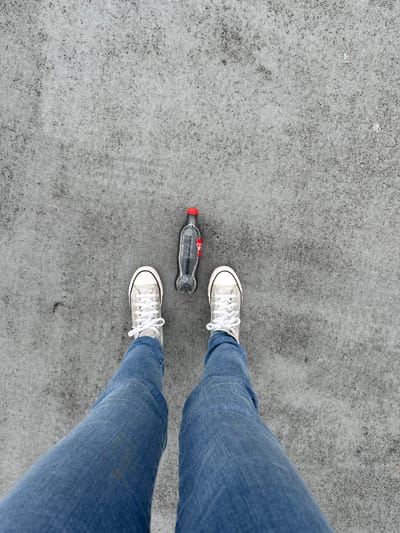 A high-angle shot shows a person wearing blue jeans and white Converse sneakers standing on a concrete surface. An empty, partially crushed Coca-Cola bottle with a red cap lies between the person's feet. The surface is predominantly gray with a textured appearance. The photo's location cannot be identified.