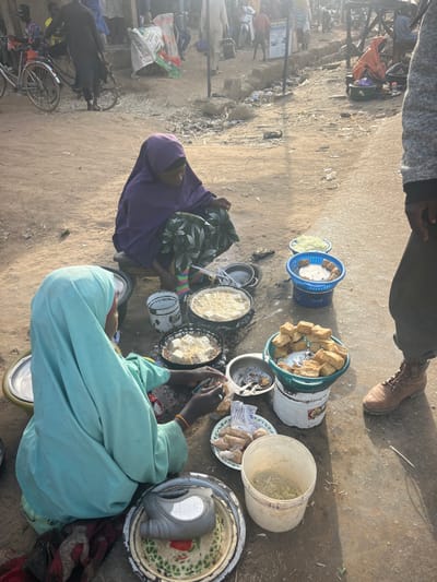Two women in traditional dress sit on the ground, preparing food in pans. Fried dough pastries are displayed in baskets and on plates. A person stands over the scene. Buckets, pots, and various containers surround them. A bicycle is to the left, and a goat is grazing nearby. The setting appears to be an outdoor market or roadside food stall.