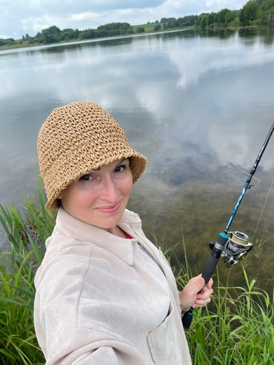 A middle-aged woman with brown hair and brown eyes, wearing a beige straw hat and a light beige corduroy shirt, is shown in a selfie. She is holding a fishing rod with a reel in her right hand and is looking at the camera with a slight smile. The woman is standing outdoors by a body of water, possibly a lake or river, with green reeds and grass in the foreground. The background shows a calm body of water reflecting the cloudy sky, with a treeline and hills in the distance. The location is identified as near Bereznik, Russia.