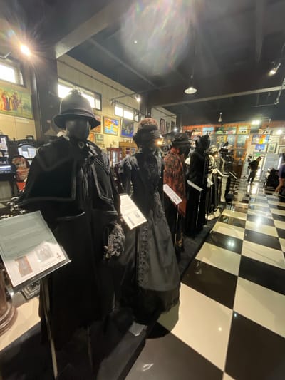 The image depicts the interior of a museum exhibition featuring mannequins dressed in historical clothing. In the foreground, a mannequin on the left wears a dark, elaborate Victorian-era outfit consisting of a coat, cape, and hat, with a descriptive placard in front of it. To its right, another mannequin is dressed in a similar dark, ornate gown. Several other mannequins in period attire are visible in a line receding into the background. The exhibition space has a black and white checkered floor. The walls are adorned with framed artwork and displays. The lighting is artificial, with visible ceiling lights and spotlights. The overall impression is of a museum showcasing historical fashion. The image is not geographically specific to Voskhod, Russia, as there are no visual cues or context provided within the image to indicate this location.