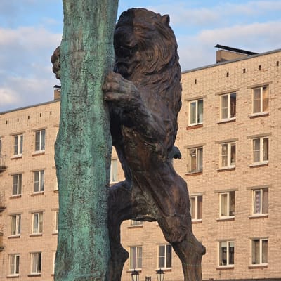 This image presents a bronze lion sculpture located in Lomonosov, Russia, observed in an urban setting. The lion, depicted with a full mane, is shown in a dynamic, upright posture, appearing to climb or cling to a thick, vertical column. The column is heavily patinated with green verdigris, contrasting with the lion's darker, reddish-brown bronze finish. Its front paws are firmly gripping the column. In the background stands an at least five-story residential building, constructed of light-colored brick. This building features multiple rectangular windows with white frames, some showing internal reflections. The building's flat roof, with some dark utility structures, is silhouetted against a blue sky dotted with scattered white clouds. The tops of two black street lamp fixtures are partially visible at the lower edge of the frame. No human figures are present in the scene.