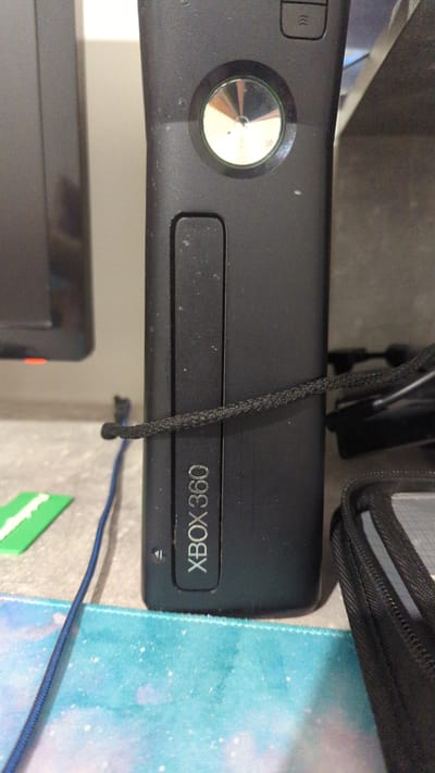 In Zadebri, Ukraine, a close-up shot captures a black Xbox 360 console, positioned vertically on a desk. The console displays the prominent "XBOX 360" branding and a circular, metallic button. A dark, braided cable is coiled around the console, securing it in place. Adjacent to the console, a black zippered case is visible, along with a bright green rectangular object and a blue cable snaking across a teal and white speckled mousepad. The background is out of focus, hinting at a dark, possibly wooden surface and a blurry television screen on the left. The lighting is dim, casting subtle shadows and highlighting the textures of the console and its accessories.