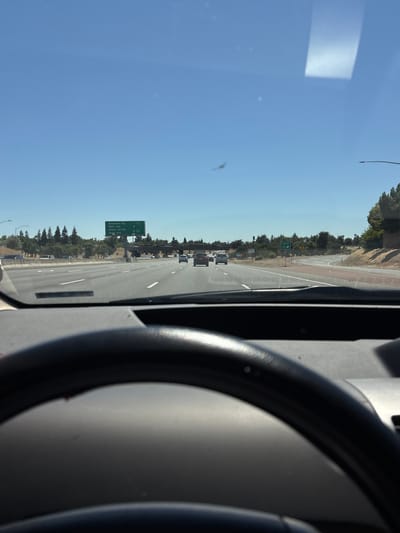 The image is a first-person perspective from the driver's seat of a car on a highway. The steering wheel is visible in the foreground, partially obscuring the view of the road. The car is traveling on a multi-lane highway under a clear blue sky. There are other vehicles on the road ahead. To the right, there is a green overhead highway sign indicating exits for Bradshaw Rd, Watt Ave, Power Inn Rd, and 29th Ave, with distances. Further ahead on the right, there is an exit sign for "EXIT 15". Trees and foliage line the sides of the highway, and an overpass is visible in the distance. The location is Rancho Cordova, California, United States of America.